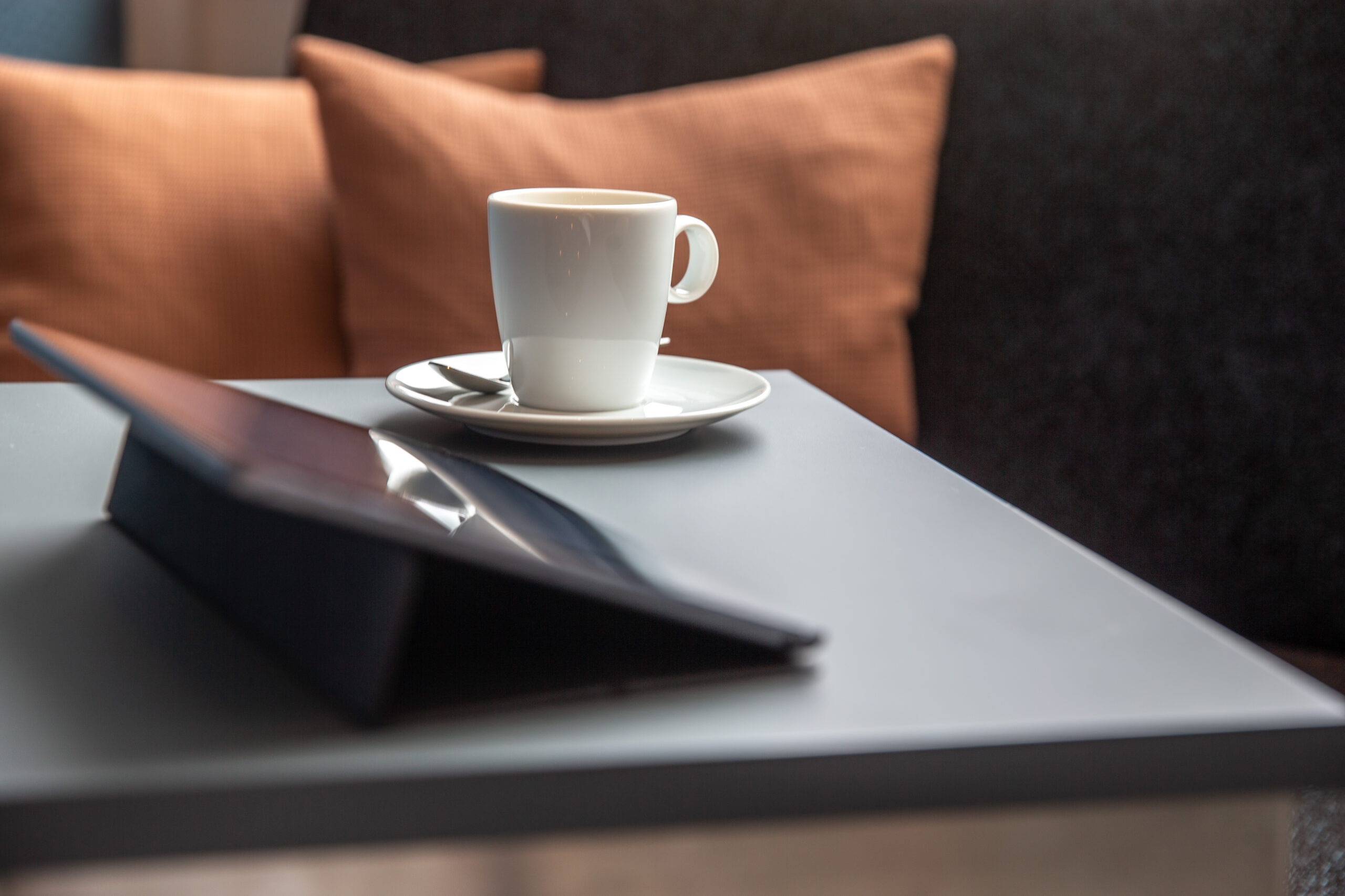 Detailansicht Kaffetasse auf Tisch mit Ipad-47 Eine weiße kaffeetasse steht auf einem nidrigen Sofatisch vor einem Ipad. Im Hintergrund sieht man ein oranges Kissen